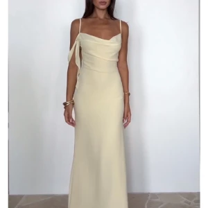 Ljus långklänning med slits och snörning - Supersnygg ljus långklänning med smala axelband, snörningsdetalj på ena axeln och öppen rygg. Klänningen har en hög slits på sidan och en elegant, figurnära passform. Perfekt för dig som vill sticka ut med en stilren och chic look.