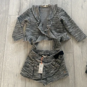 Gråmelerade shorts med rosett Mini A Ture - Super Snygga gråmelerade shorts från Mini A Ture med tillhörande kofta  i stickad look. Jättesöt på flickor i st 3-5 årDe har hög midja, dekorativ svart rosett och ett litet metallmärke framtill. Materialet är mjukt och strukturerat, perfekt för en trendig och bekväm stil.