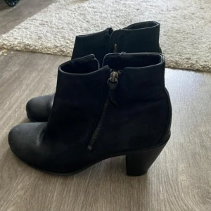 Svarta stövletter från Ecco i skinn - Snygga svarta stövletter från Ecco i mjukt skinn med dragkedja på sidan och stabil klack. Skorna har en klassisk rund tå och platt sula undertill, vilket gör dem både stilrena och bekväma. Perfekta för dig som gillar enkel och tidlös design. 