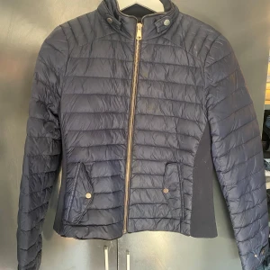 Mörkblå dunjacka från Zara Woman - Snygg mörkblå dunjacka från Zara Woman med quiltad design och dragkedja framtill. Jackan har ståkrage med tryckknappar, två fickor med knappstängning och smidiga paneler i sidorna för extra komfort. Perfekt för kyliga dagar och enkel att matcha med allt.