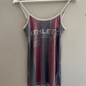 Randig linne-topp med tryck - Säljer en sportig linne-topp med smala axelband och trycket 'ATHLETIC 07' framtill. Toppen har breda vertikala ränder i vinrött och grått, med ljusgrå kant runt hals och ärmhål. 