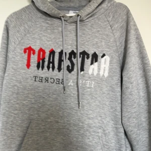 Grå hoodie från  med tryck - Säljer en grå hoodie från Trapstar med fet logga i rött, svart och vitt på bröstet. Klassisk känguruficka, snörning i huvan och mjukt material. Perfekt för dig som gillar streetwear och vill sticka ut med en ikonisk look.