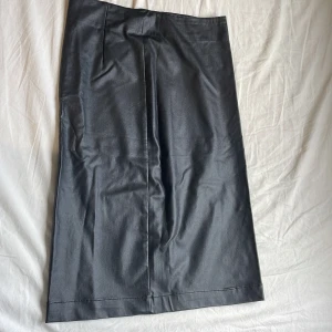 Svart långkjol i skinnlook, stl 36 - Snygg svart långkjol från First Class Collection i skinnimitation. Kjolen har rak passform, diskret slits bak och dragkedja i sidan för enkel påtagning. Perfekt för dig som gillar en stilren och edgy vibe.