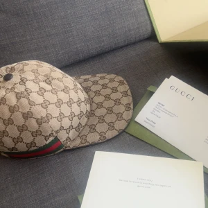 Gucci keps med GG-mönster och rödgrön detalj - Snygg keps från Gucci i beige och brunt med klassiskt GG-monogram över hela kepsen. Kepsen har justerbar rem bak i svart skinn och en ikonisk rödgrön rand på sidan. Perfekt accessoar för dig som vill sticka ut med lyxig streetstyle. Kan även bytas!