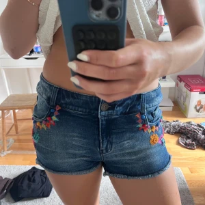 Blå jeansshorts med broderade blommor - Snygga blå jeansshorts med färgglada broderade blommor och mönster framtill och baktill. Shortsen har råa kanter nertill och klassisk femficksmodell. Perfekta för sommaren och ger en cool, bohemisk vibe med sina detaljerade broderier.