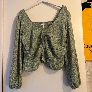 Grön blommig blus från H&M - Supersöt grön blus från H&M med små vita blommor. Blusen har långa puffärmar med resår vid handlederna, v-ringning och knyt framtill. Smockad rygg och volangkant nedtill ger en extra gullig touch. Perfekt för vår och sommar! Blusen är i ett mycket fint skick.