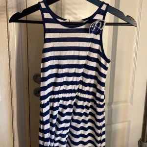 Randig långklänning  - Långklänning med marinblå och vita horisontella ränder, ärmlös design och rund halsringning. Klänningen har en söt blomdetalj vid axeln och resår i midjan för en avslappnad passform. Perfekt för varma dagar och enkel att styla.