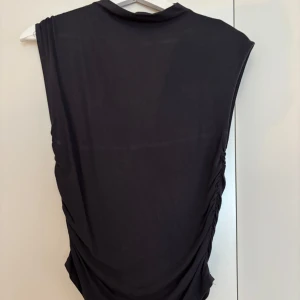 Svart topp från Gina Tricot - Svart topp från Gina Tricot soft touch med ärmlös design och rynkade sidor. Klänningen har en enkel, stilren look med hög hals och är gjord i mjukt bomullsmaterial. Perfekt för dig som gillar minimalistisk stil och vill ha något bekvämt men snyggt.