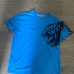 Blå t-shirt med tigertryck - Cool blå t-shirt med svart tigertryck över axeln. Klassisk passform med rund hals och korta ärmar. Perfekt för dig som vill sticka ut med ett djurmotiv. Materialet känns mjukt och lätt, perfekt för varma dagar.