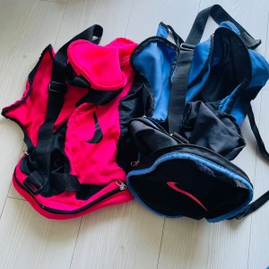 Nike träningsväskor rosa & blå - Två sportiga träningsväskor från Nike, en i stark rosa med svarta detaljer och en i blått och svart. Båda har justerbara axelremmar, dragkedjor och den klassiska Nike-loggan på sidan. Perfekta för gymmet eller sportaktiviteter.
