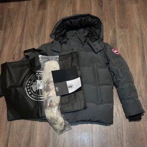 Canada Goose jacka  - Säljer en grå jacka från Canada Goose i mycket bra skick. Jackan har en avtagbar huva och är perfekt för kalla vinterdagar. Den har dragkedja och knappar framtill samt två stora fickor. Märkets logga syns på armen. Supervarm och oöppnad päls medföljer! Pris kan sänkas hör av er.