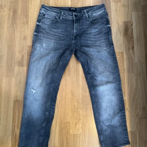 Jack&Jones jeans - Säljer ett par snygga grå jeans från Jack & Jones med slitningar och tvättad look. Klassisk femficksmodell med dragkedja och knapp. Jeansen har raka ben och normal passform, perfekta till en avslappnad stil.