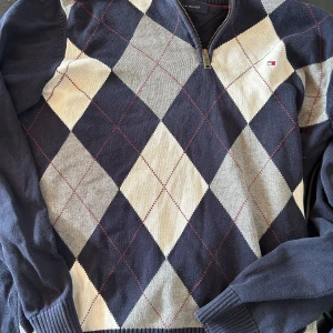 Argyle half zip tröja Tommy Hilfiger - Snygg stickad tröja från Tommy Hilfiger med klassiskt argylemönster i marinblått, grått och vitt. Tröjan har half zip-dragkedja, långärmad passform och diskret logga på bröstet. Perfekt för dig som gillar preppy stil.