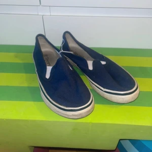 Mörkblå slip-on sneakers Sport strl 34 - Mörkblå slip-on sneakers från Sport i storlek 34. Skorna har vit sula med svart rand och är tillverkade i textil. Klassisk och enkel design med rund tå och elastiska partier vid öppningen för enkel påtagning.