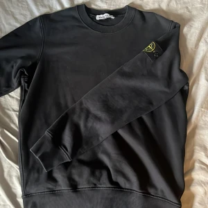 Svart sweatshirt från Stone Island - Svart crewneck sweatshirt från Stone Island med klassisk logotyp-patch på vänster ärm. Tröjan har rund halsringning, ribbade muddar och är tillverkad i mjuk bomull. Perfekt för dig som gillar streetwear och vill ha en clean look med ikonisk detalj. (Inga defekter men har använts hyfsat mycket)