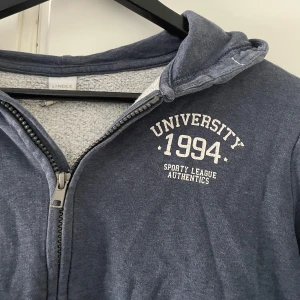 Blå hoodie med dragkedja från Lindex - Mörkblå hoodie från Lindex med vit text 'University 1994 Sporty League Authentics' på bröstet. Tröjan har huva, dragkedja framtill och känguruficka. Tillverkad i mjuk bomullsmix, perfekt för chill dagar.