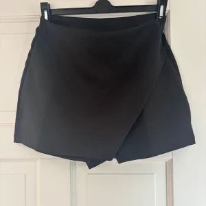 Svart skort i kort modell - Svart kort omlottkjol med stilren design och diskreta fickdetaljer baktill. Kjolen har en modern passform och är perfekt för dig som gillar minimalistisk stil. Tillverkad i ett mjukt och följsamt material som känns bekvämt hela dagen.