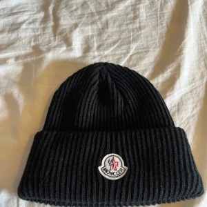 Svart Moncler mössa i ribbstickad design - Säljer en svart ribbstickad mössa från Moncler med den klassiska loggan på framsidan. Mönstret är tydligt med breda ribbor som ger en varm och stilren look. Perfekt för kalla dagar, tillverkad i mjukt och värmande material. En snygg accessoar som kombinerar funktion och mode.