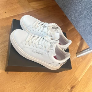 Vita Love Moschino sneakers med hjärtdetalj - Snygga vita sneakers från Love Moschino med silvriga hjärtdetaljer bak och logga på sidan. Skorna har snörning, platt sula med hjärtmönster undertill och röda hjärtan i innersulan. Perfekta för dig som gillar stilrena men lekfulla detaljer.