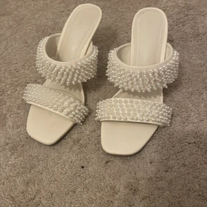 Vita sandaler med pärldetaljer och klack - Unika vita sandaler med två breda remmar täckta av pärlor för en extra glam känsla. Skorna har fyrkantig tå och låg klack, perfekt för dig som vill sticka ut. Tillverkade i syntetmaterial och har öppen tå. Snygga till alla outfits när du vill ha något extra. Storlek 37 men skulle kunna passa 36 rätt bra