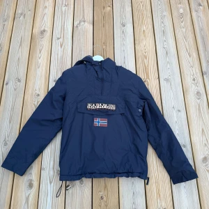 Blå vindjacka från Napapijri - Snygg marinblå vindjacka från Napapijri med halv dragkedja och stor ficka framtill. Jackan har huva, lång ärm och norsk flagga som detalj på bröstet. Perfekt för blåsiga dagar och streetwear-stil.
