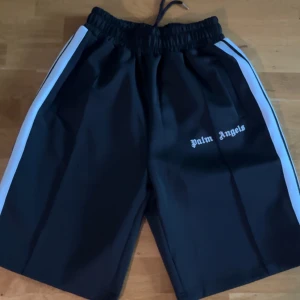Palm Angels svarta shorts med vita ränder - Svarta shorts från Palm Angels med vita ränder längs sidorna och logga tryckt på vänster ben. Modellen har elastisk midja med snörning och sportig vibe. Perfekta för dig som gillar streetwear och vill sticka ut med en clean look. Shortsen är storlek M men är stora i storleken.