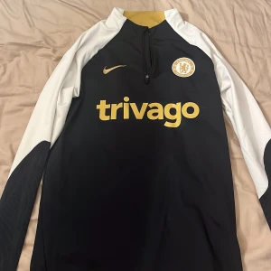 Chelsea långärmad Nike Dri-Fit tröja S - Chelsea FC långärmad träningströja från Nike i svart och vitt med guldiga detaljer. Tröjan har broderat klubbmärke, Nike-logga och Trivago-tryck på bröstet. Tillverkad i Dri-Fit-material som andas och har en slim fit-passform. Perfekt för fotbollsträning eller support. Köpt i Chelseas butik i London. Knappt använd, mycket bra skick!