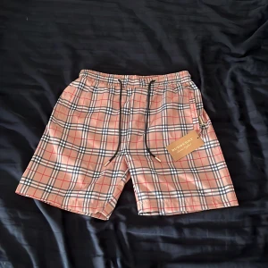Burberry shorts  - Snygga shorts från Burberry i klassiskt beige, svart, vit och röd rutig design. De har elastisk midja med svart snörning och är tillverkade i ett lätt och bekvämt material, perfekt för sommaren. Ikonisk look som sticker ut och passar dig som gillar exklusiva märken.