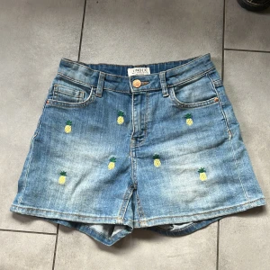 Jeansshorts från Lindex i storlek 146 - Blå jeansshorts från Lindex med broderade små ananaser. Fickor bak och fram, knappstängning och justerbar midja. Använda men i bra skick, inga tydliga märken eller defekter.
