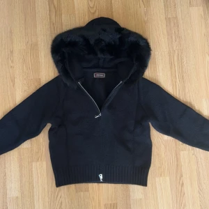 Alessa Fur Jacket  - Mysig svart stickad jacka från Alessa med huva kantad av fluffig  päls. Jackan har dragkedja framtill och breda ribbade muddar vid ärmslut och nederkant. Perfekt för kalla dagar när du vill vara både varm och snygg. Helt nya aldrig använd 
