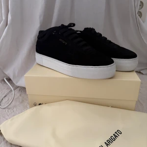 Svarta sneakers från Axel Arigato - Stilrena svarta sneakers från Axel Arigato i mocka med vit platt sula. Klassisk låg modell med snörning och diskret guldfärgad logga på sidan. Perfekta för dig som gillar minimalistisk och clean stil.