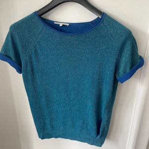 Blå glittrig Maje- topp med kort ärm - Snygg blå glittrig topp med korta ärmar från Maje. Toppen har en lätt glansig finish och uppvikta ärmslut i en något mörkare blå nyans. Har fått många komplimanger för den och är perfekt både till vardags och fest.