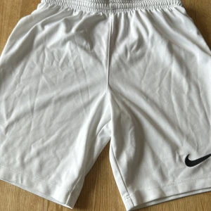 Vita Nike Dri-Fit shorts - Snygga vita sportshorts från Nike med Dri-Fit-teknologi som håller dig torr. De har elastisk midja och en klassisk svart Nike-logga på benet. Perfekta för träning eller fotboll, tillverkade i lätt och snabbtorkande polyester.