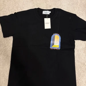 Svart Casablanca t-shirt med tryck - Svart t-shirt från Casablanca med färgglatt grafiskt tryck i blått, gult och vitt både fram och bak. Klassisk passform med rund hals och korta ärmar. Perfekt för dig som gillar stilrena plagg med en twist. Materialet känns mjukt och skönt mot huden.
