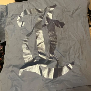 Blå t-shirt med silvertryck Juicy Couture - Snygg ljusblå t-shirt från Juicy Couture med ett stort metallic silvertryck på framsidan. Klassisk passform och korta ärmar, tillverkad i mjuk bomull. Perfekt för dig som vill sticka ut med en cool och trendig look.