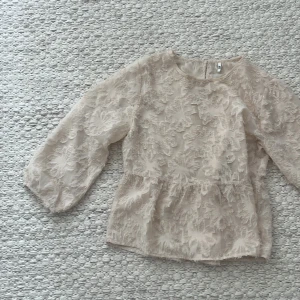 Beige blus med blommig struktur från NLY - Supersöt beige blus från JDY med transparent tyg och stora blommiga strukturer över hela toppen. Blusen har rund halsringning och vida ärmar som ger en romantisk vibe. Perfekt för dig som gillar detaljer och en feminin look.