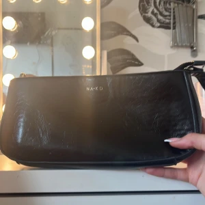 Svart axelväska från NA-KD i skinn - Snygg svart axelväska från NA-KD i glansigt skinnmaterial. Väskan har en stilren, avlång form med guldfärgad dragkedja och justerbart axelband. Perfekt för att bära det viktigaste och ger en clean look till din outfit.
