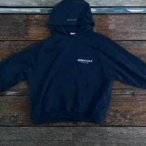 Mörkblå Essentials hoodie Fear of God - Snygg mörkblå hoodie från Fear of God Essentials med vit logga på bröstet och ärmen. Klassisk känguruficka, huva och relaxed fit. Perfekt för dig som gillar streetwear och vill ha en stilren look. Materialet är mjuk bomullsmix som känns skön mot huden.
