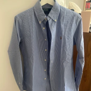 Blårutig skjorta från Ralph Lauren - Säljer en stilren blårutig skjorta från Ralph Lauren med broderad logga på bröstet. Skjortan har långa ärmar och knappar framtill samt på kragen. Perfekt för en klassisk slimfit look. Storlek S men funkar för S.
