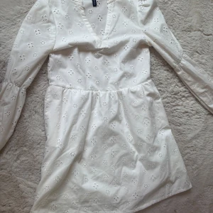 Vit broderad kortklänning med v-ringning - Superfin vit kortklänning från H&M med broderade blommor och v-ringning. Klänningen har långa ärmar och är gjord i bomull, perfekt för vår och sommar. Söt och fräsch stil som passar till många tillfällen.