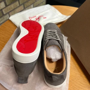 Gråa sneakers från Christian Louboutin - Säljer ett par gråa sneakers från Christian Louboutin med klassisk röd sula. Skorna har snörning, rund tå och är tillverkade i mocka med vita gummisulor. Perfekta för dig som gillar stilrena och exklusiva sneakers. Storlek 42 men passar 41