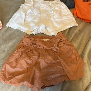 Två par shorts med knytband - Säljer två par snygga shorts, ett par vita och ett par rostbruna. Båda har hög midja, bred resår och knytband i midjan för extra stilpoäng. Stora fickor framtill och mjukt, lätt material som passar perfekt till varma dagar.
