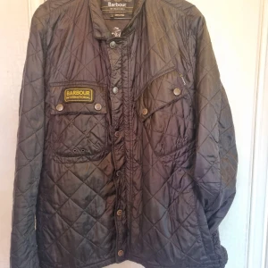Svart quiltad jacka Barbour L - Quiltad svart jacka från Barbour International med coolt motorcykeltryck på insidan. Jackan har tryckknappar, två bröstfickor med lock och Barbour-märke på vänster sida. Perfekt för dig som gillar klassisk brittisk stil och snygga detaljer.