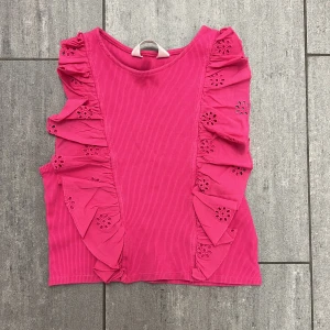 Rosa ribbad linnetopp från H&M - Supersöt rosa linnetopp från H&M med ribbad struktur och volangdetaljer längs sidorna. Volangerna har broderade blommor som ger toppen en extra charmig touch. Perfekt för dig som vill sticka ut med färg och detaljer.