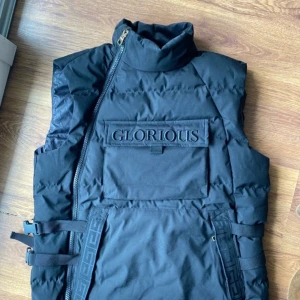 Svart dunväst med dragkedja från Glorious - Snygg svart dunväst från Glorious med en stor ficka framtill och justerbara spännen på sidorna. Västen har en hög krage och en diagonal dragkedja för en modern look. Perfekt för kyligare dagar.