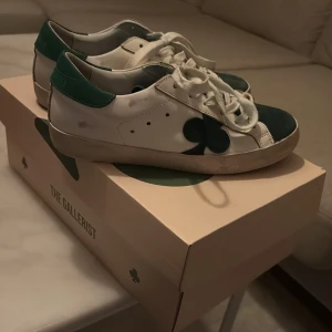 Vita och gröna sneakers från The Gallerist - Snygga vita sneakers med gröna detaljer från The Gallerist. Skorna har en unik design med gröna klöver på sidorna och snörning framtill. Perfekta för en stilren look.