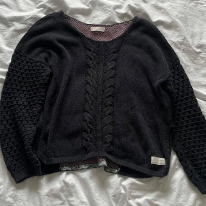 Svart stickad tröja med mönster från oddmolly - Säljer en svart stickad tröja från odd Molly med ett unikt mönster i rosa och grått på baksidan. Tröjan har långa ärmar med ett flätat mönster och en rund halsringning. Perfekt för att hålla sig varm och stilren.