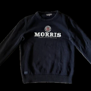 Mörkblå tröja från Morris - Säljer en stilren mörkblå tröja från Morris med text och emblem på bröstet. Tröjan har långa ärmar och ribbade muddar vid ärmslut och nederkant. Perfekt för en klassisk look.