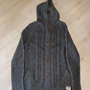 Grå stickad hoodie från Jack & Jones - Säljer en grå stickad hoodie från Jack & Jones Originals. Den har en mysig huva och knappar framtill. Perfekt för kyliga dagar med sin stilrena design och praktiska fickor. Passar både till vardags och mer avslappnade tillfällen.