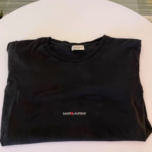 Svart t-shirt från Saint Laurent - Snygg svart t-shirt från Saint Laurent med deras ikoniska logga tryckt på bröstet. Perfekt för en stilren look. Gjord i mjukt material för extra komfort.